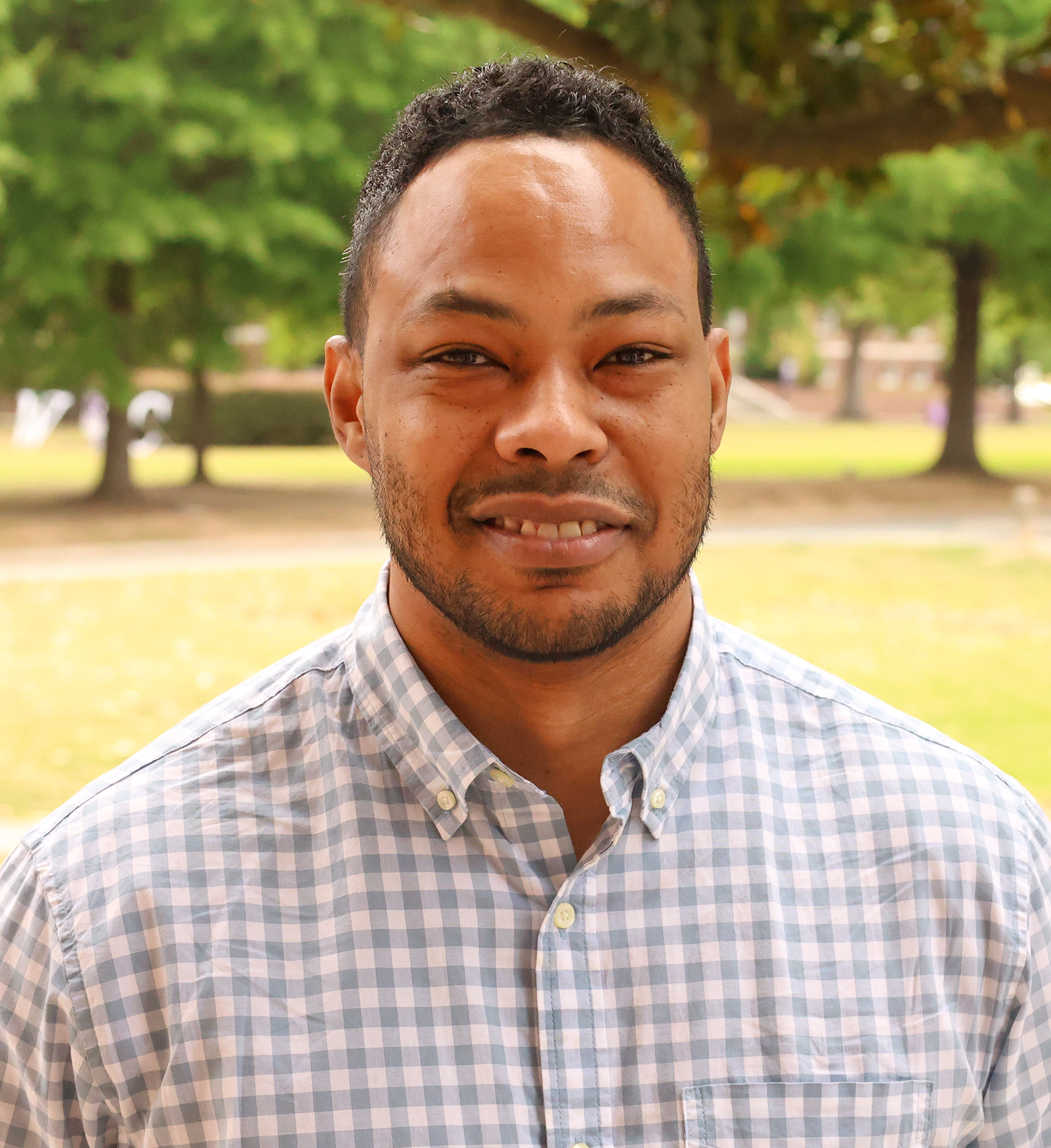 Smiling man with short curly hair and light facial hair standing outdoors on a campus lawn. He is wearing a light blue checkered button-down shirt, with trees and greenery softly blurred in the background.