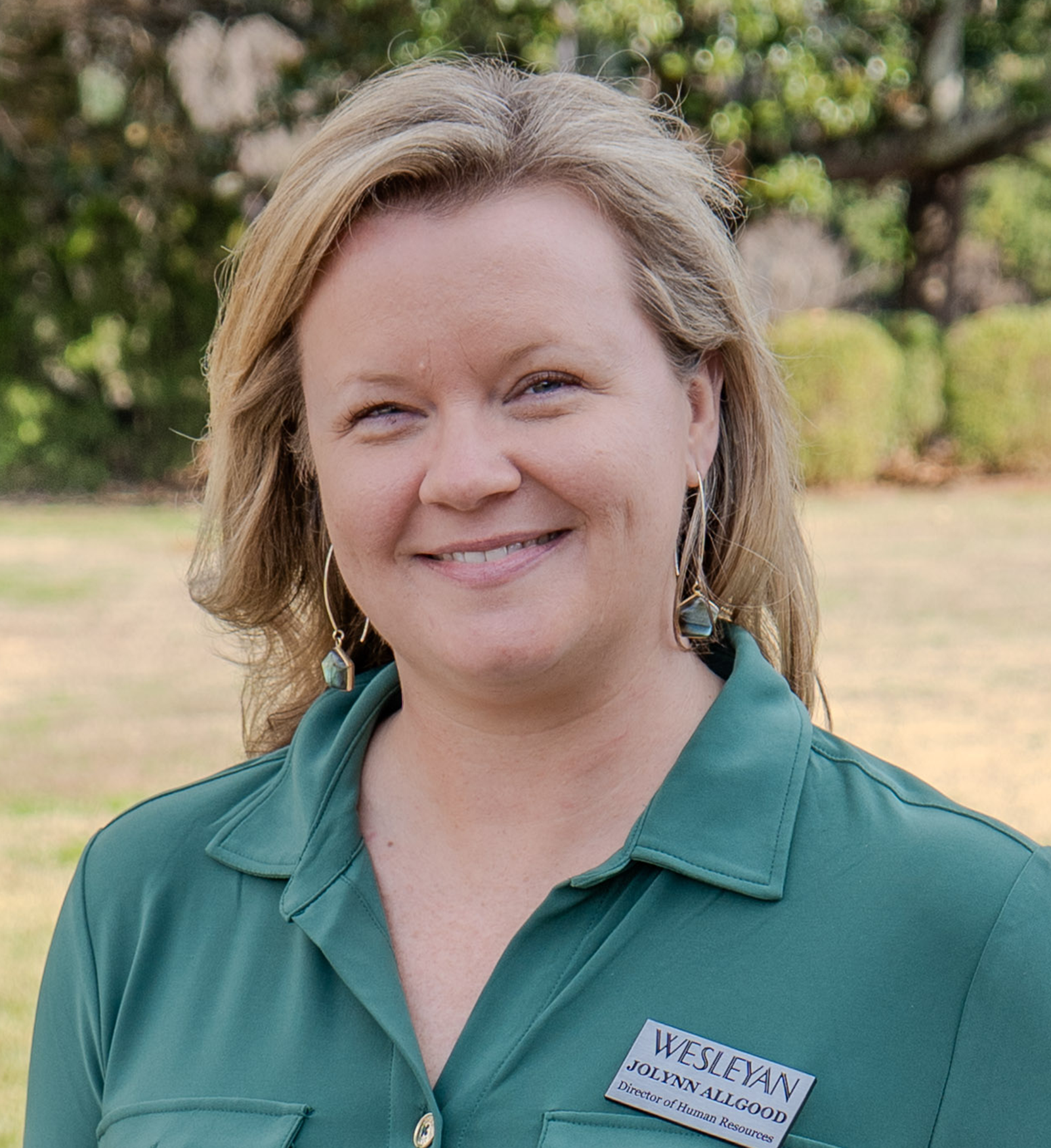 Smiling woman with shoulder-length blonde hair wearing a green collared shirt and geometric earrings, standing outdoors with trees and greenery in the background. She has a name badge that reads “Jolynn Allgood, Director of Human Resources.”