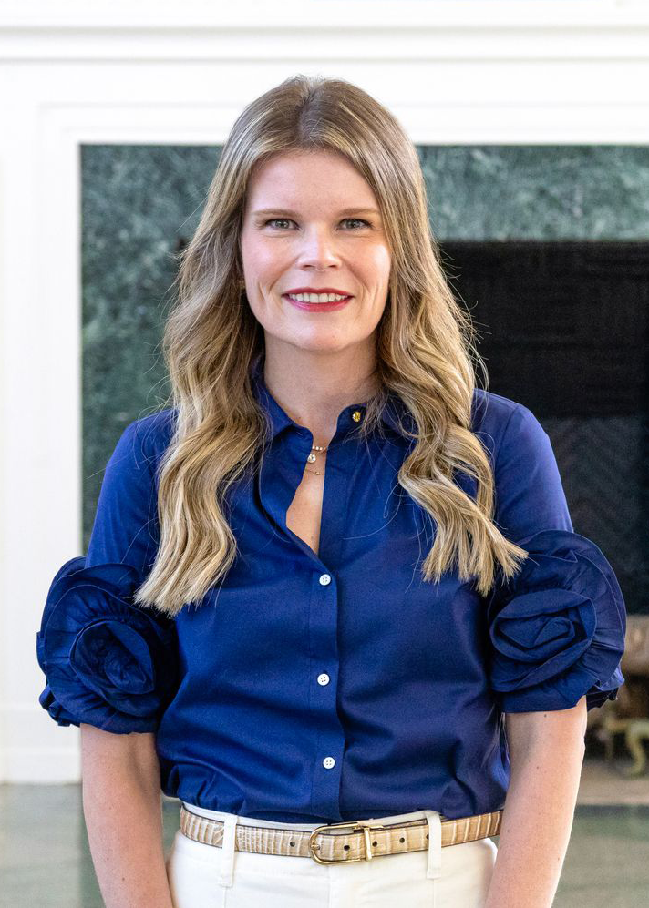 Woman with long wavy blonde hair wearing a royal blue blouse with puff sleeves and white pants, standing indoors and smiling at the camera. A fireplace and light-colored interior details are visible in the background.